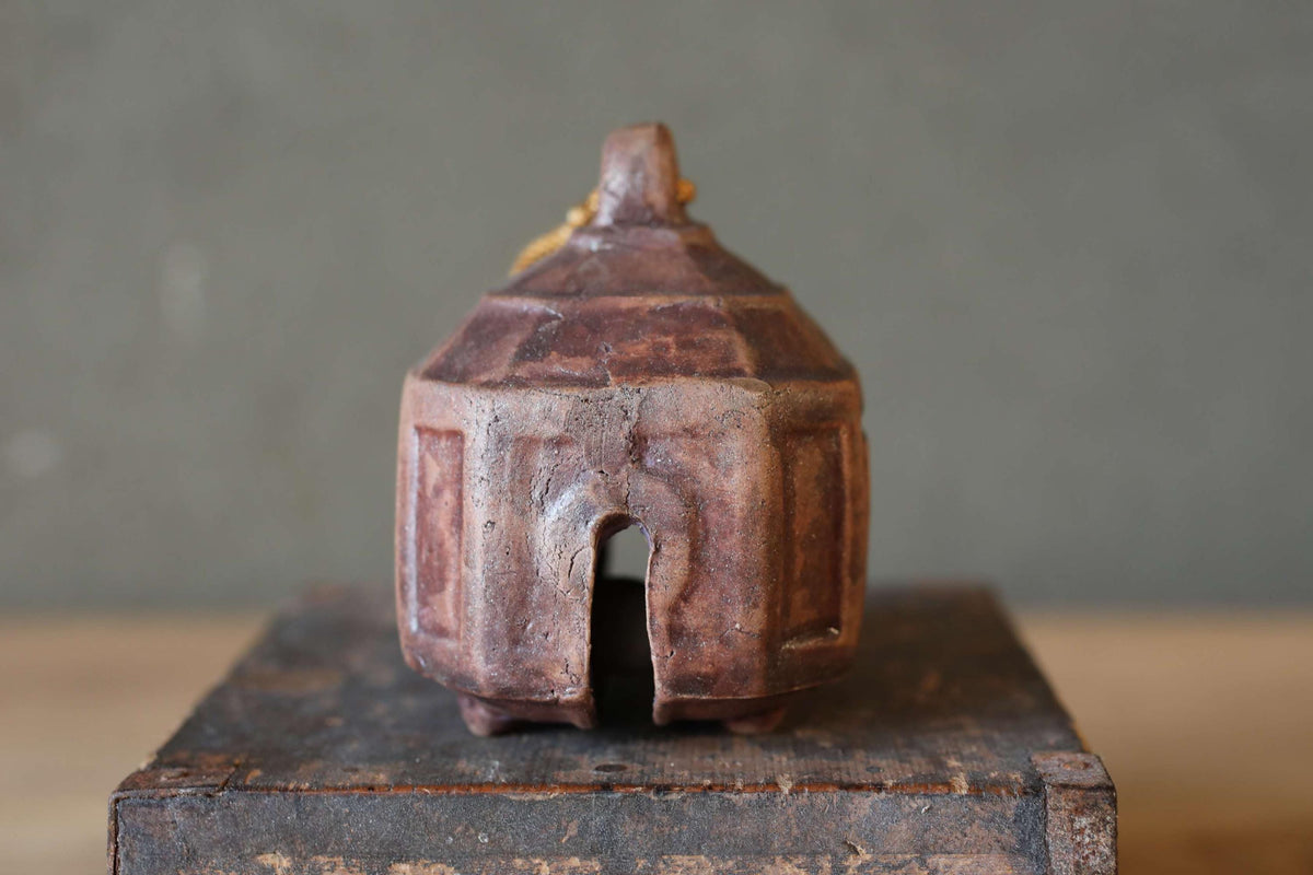 A small, brown ceramic bell or container with a pointed top sits on a wooden surface against a gray background.