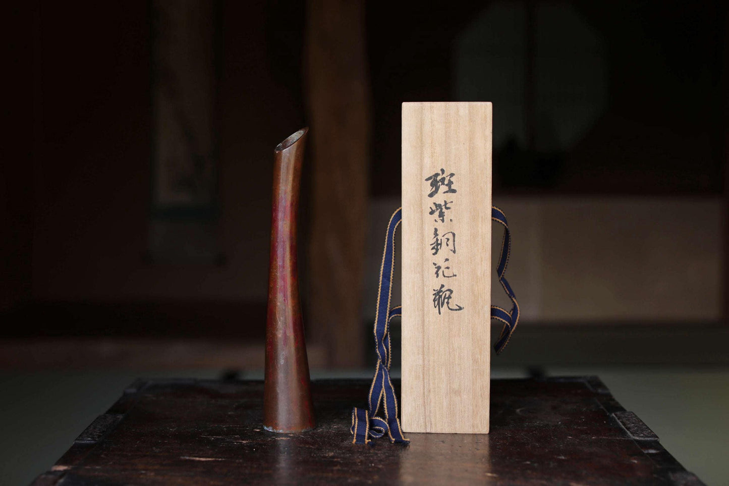 a wooden box with Japanese characters on it, placed on a dark surface, next to a tall, narrow object with a reddish-brown color.