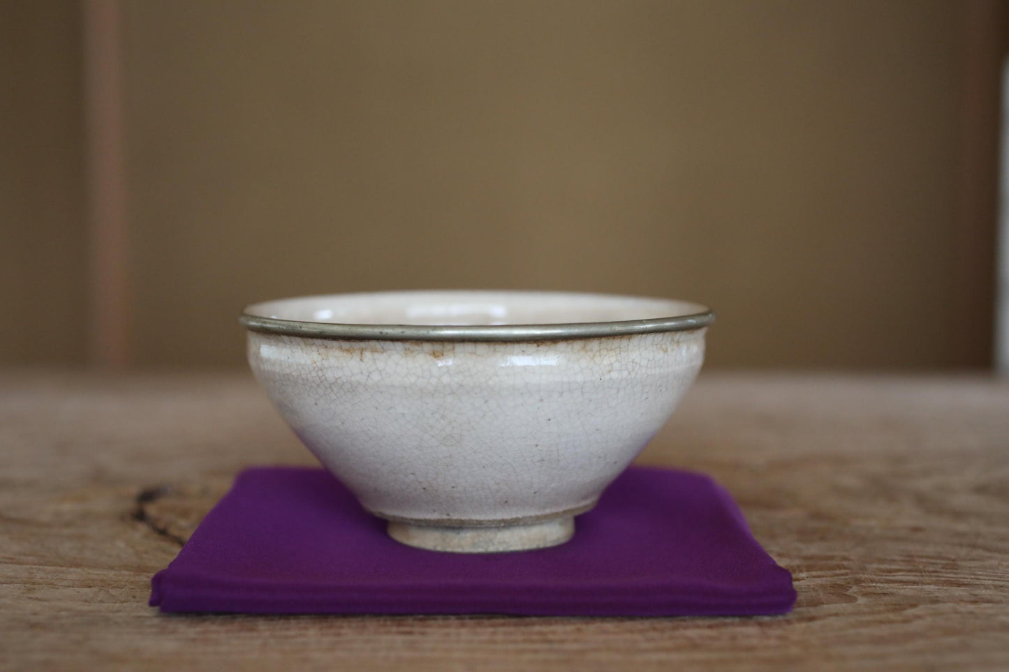 A white ceramic bowl with a silver rim sits on a purple cloth on a wooden surface.