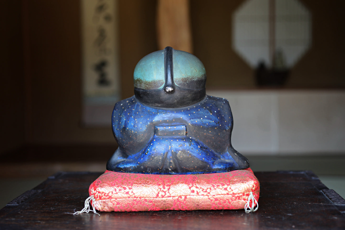 A ceramic figurine of a person wearing a blue and black outfit, sitting on a red cushion, placed on a wooden surface.