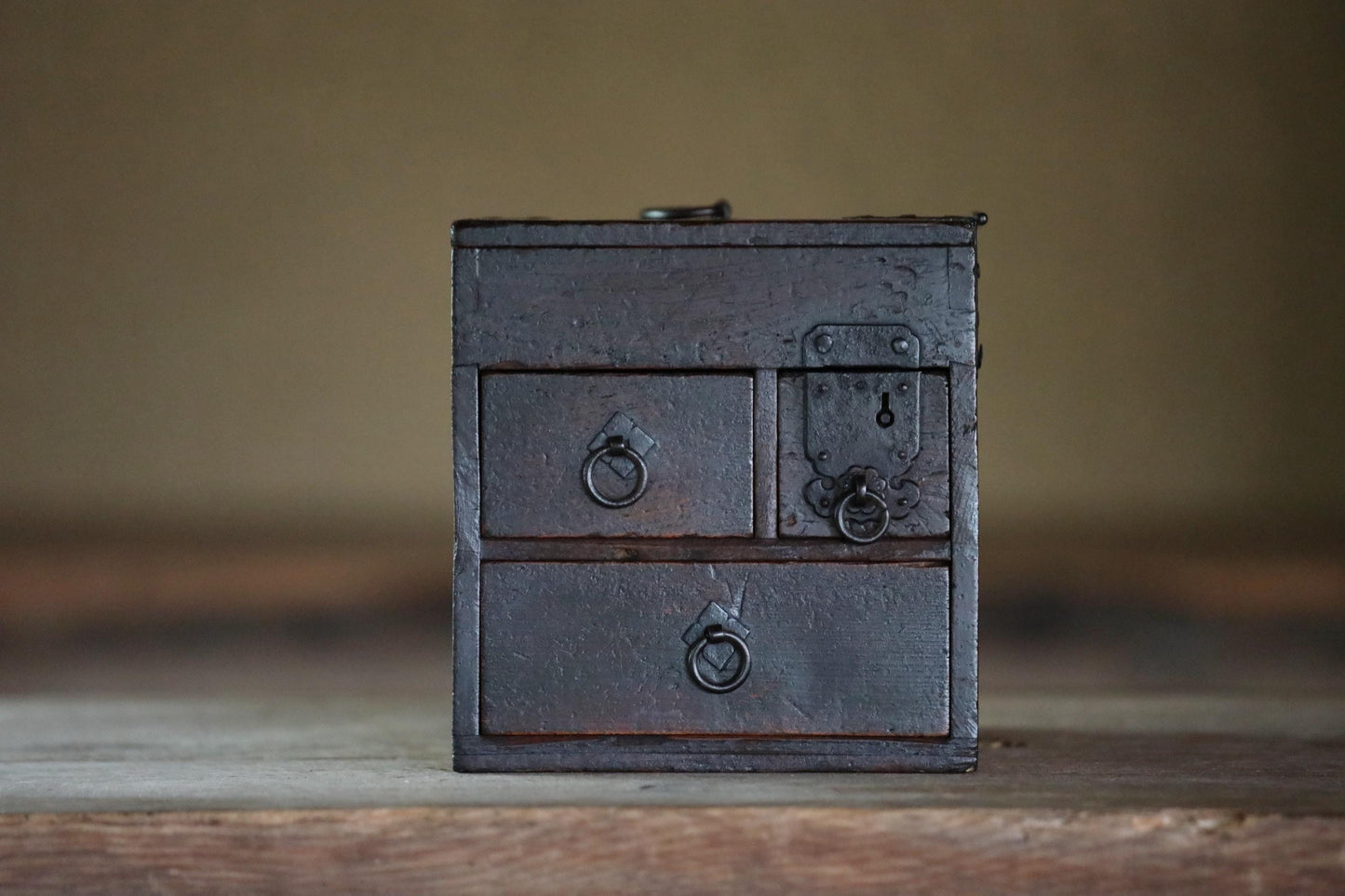 A small, old-fashioned wooden chest with three drawers sits on a wooden surface.