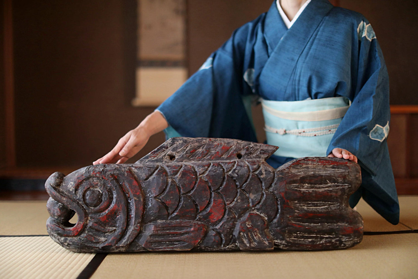A person wearing a traditional Japanese kimono is holding a large, intricately carved wooden fish.