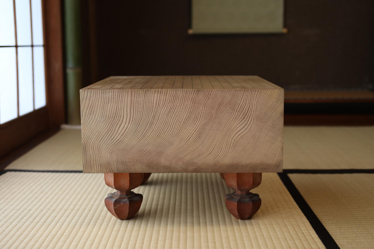 A wooden table with a natural wood grain texture and four dark-colored wooden legs, placed on a tatami mat in a room with a dark background.