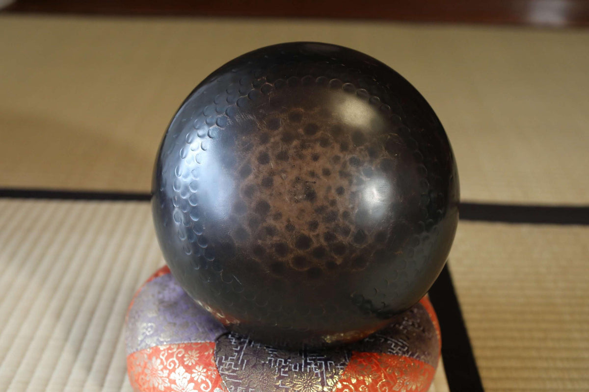 A black, textured bowl sits on a colorful, patterned cushion on a wooden surface.