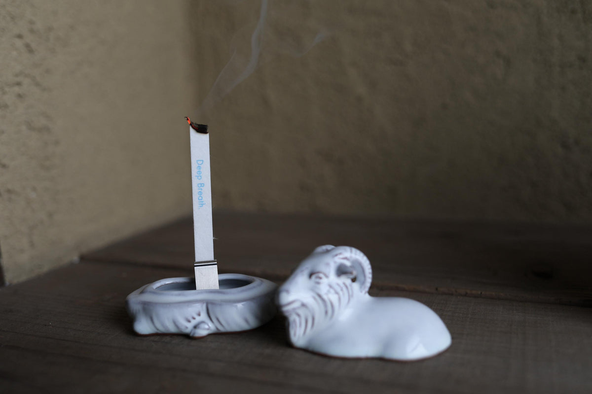A white ceramic incense burner with a lit incense stick sits on a wooden surface, with smoke rising from the incense.