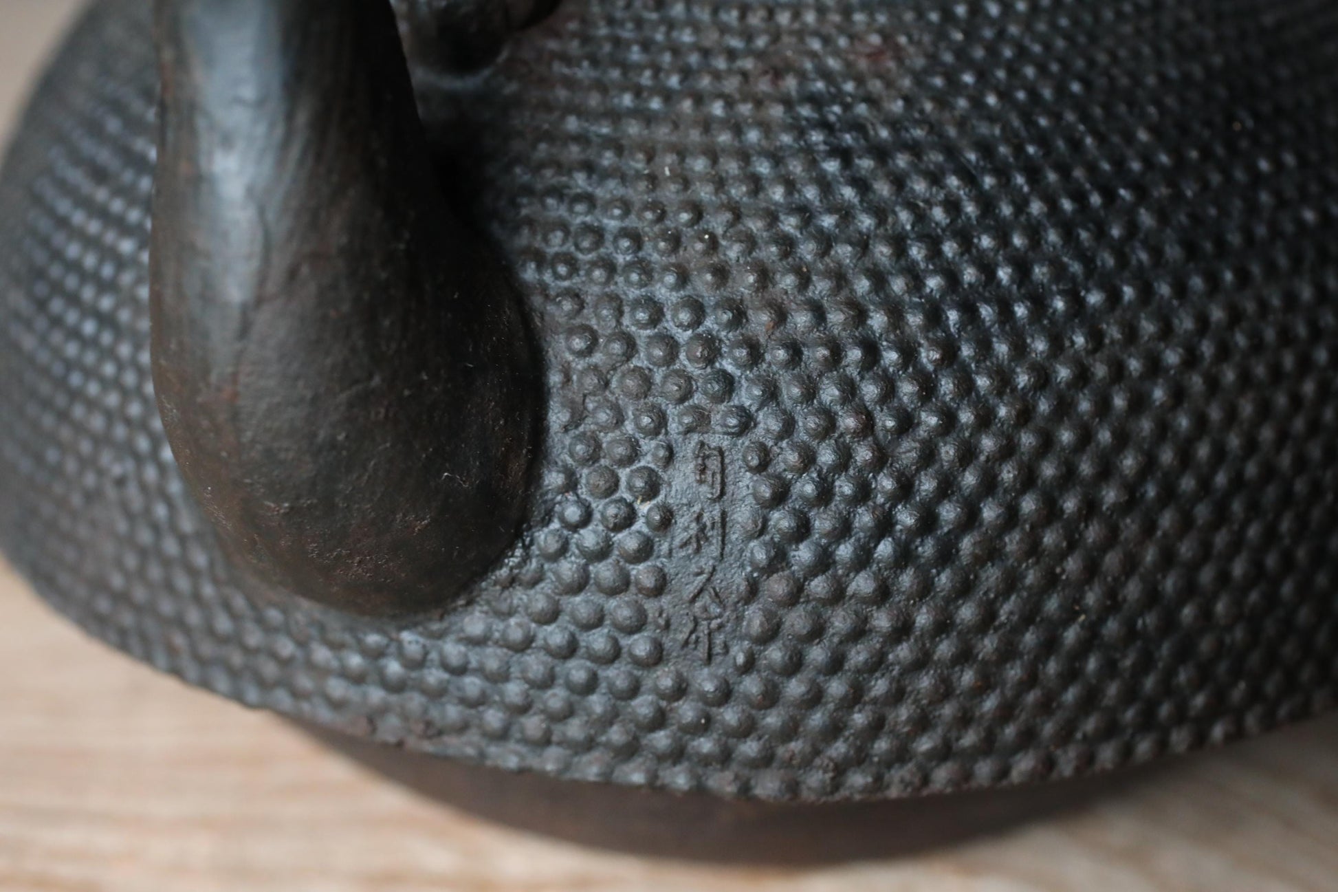 A close-up view of a black metal teapot with a textured surface, featuring a small engraved mark or signature.
