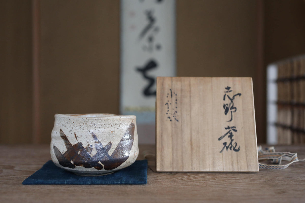a wooden box with Japanese characters on it, placed on a wooden surface next to a white ceramic bowl with a brown design.