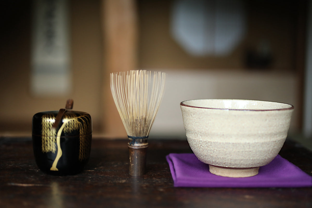 a wooden table with a black and gold container, a white bowl, and a white whisk on a purple cloth.