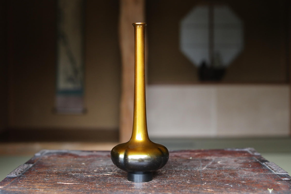 A golden vase with a long, slender shape sits on a wooden surface, with a blurred background suggesting an indoor setting.