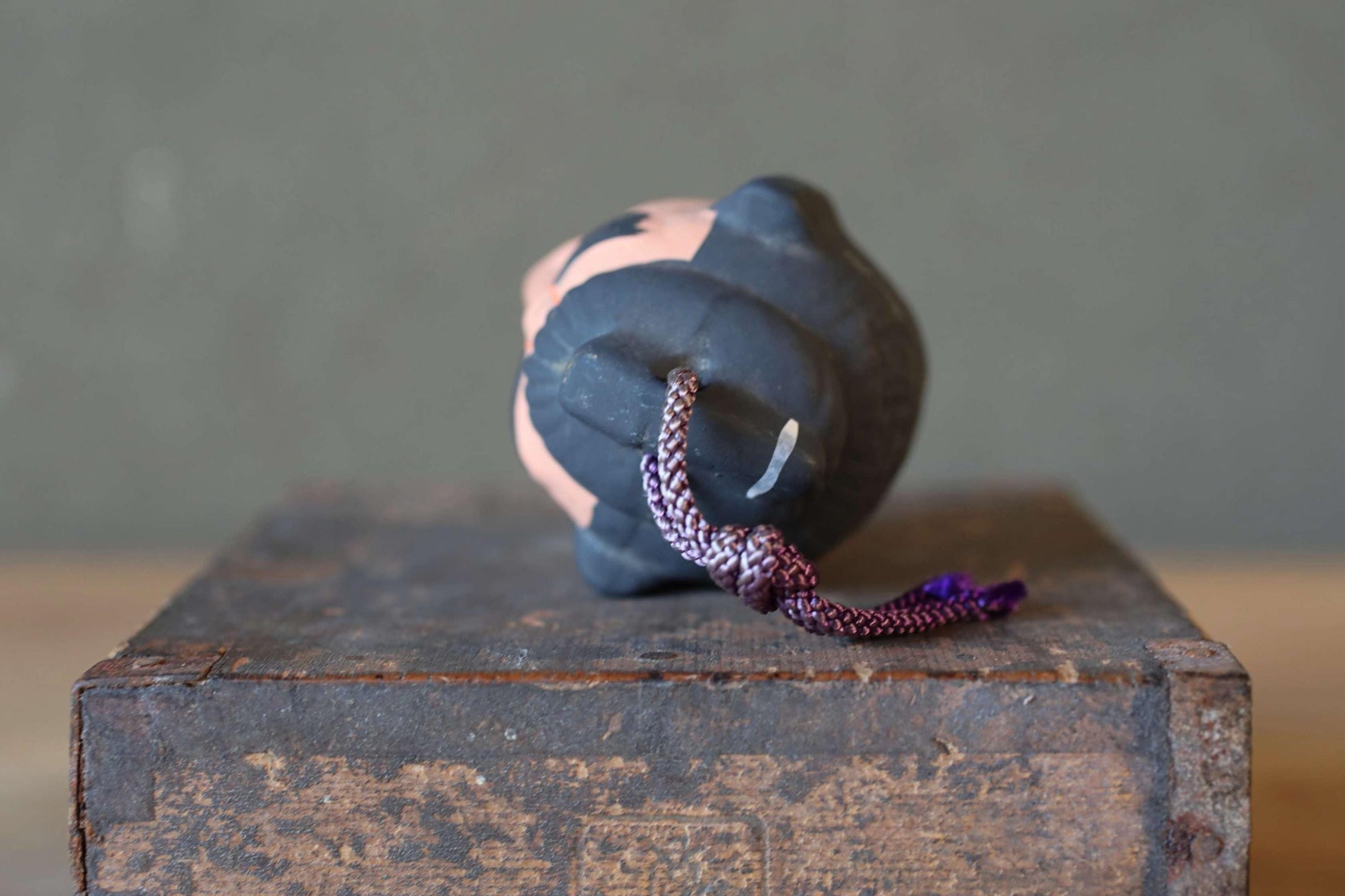 A ceramic figurine with a black and pink color scheme sits on a wooden surface, with a purple string or cord hanging from it.