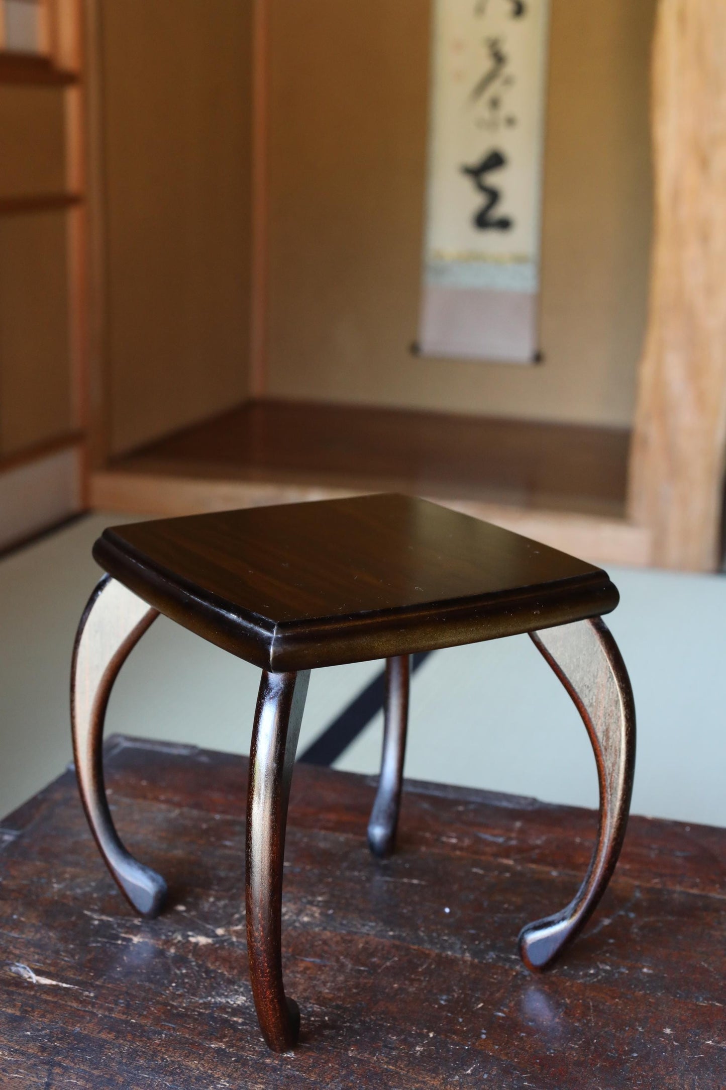 A dark wooden stool with curved legs sits on a wooden floor in a room with a wooden wall and a hanging paper with Japanese characters.