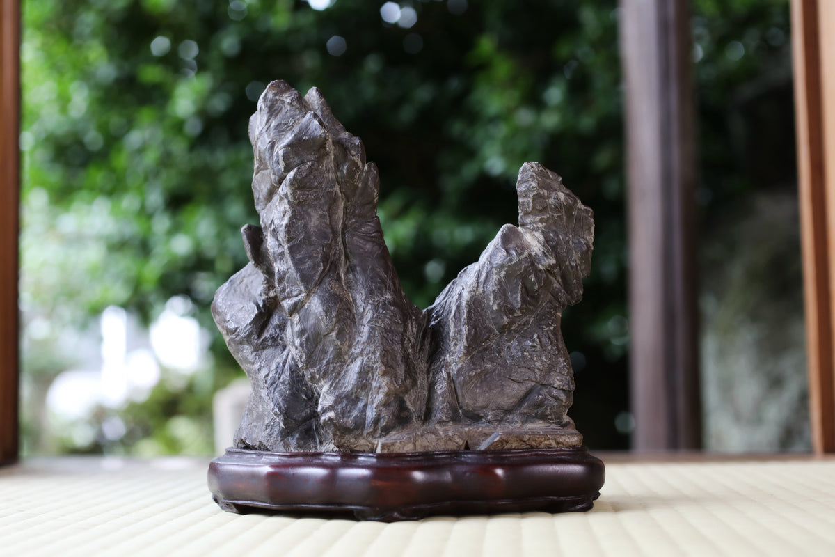 A stone sculpture of two large rocks sits on a wooden base, placed on a table with a blurred natural background.
