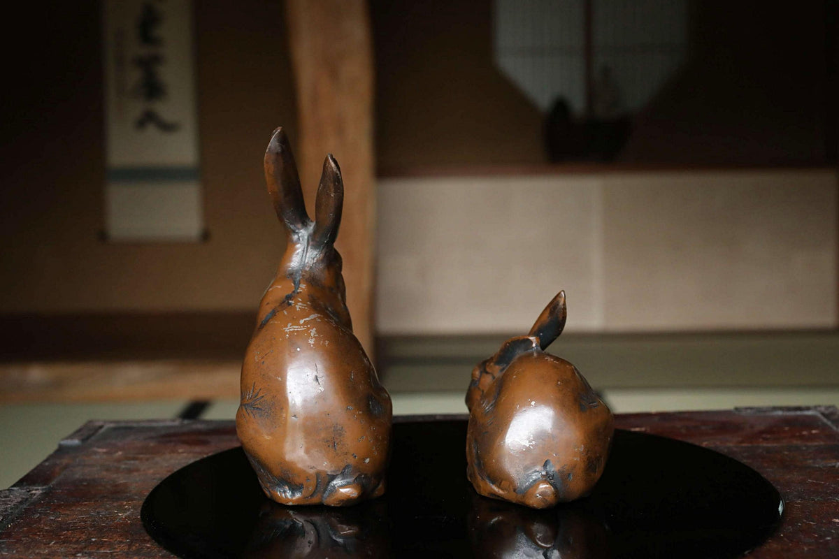 Two bronze rabbit figurines are displayed on a black circular surface, with a blurred background suggesting an indoor setting.