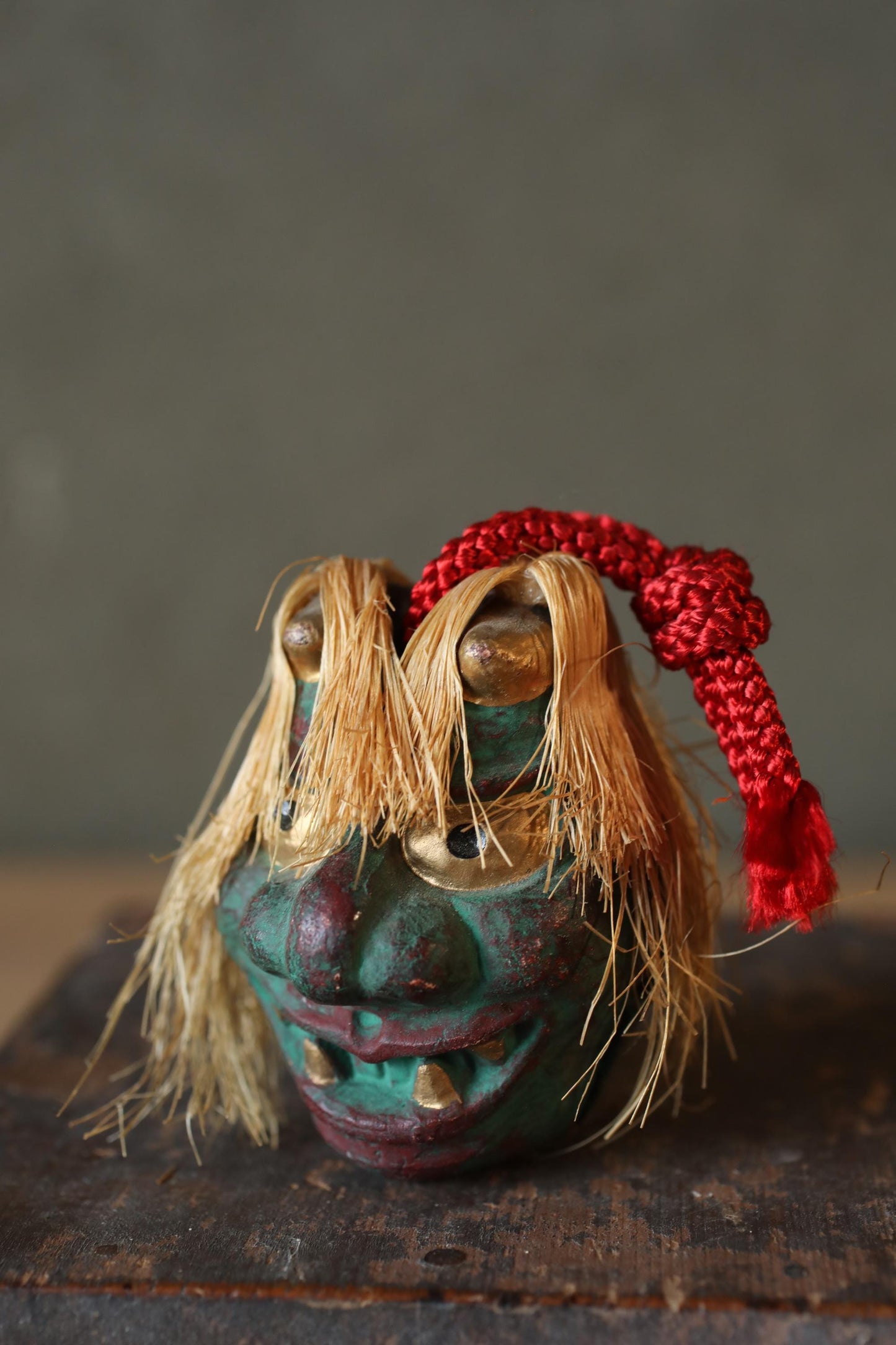 A colorful, whimsical mask with a red ribbon around its neck, sitting on a wooden surface.
