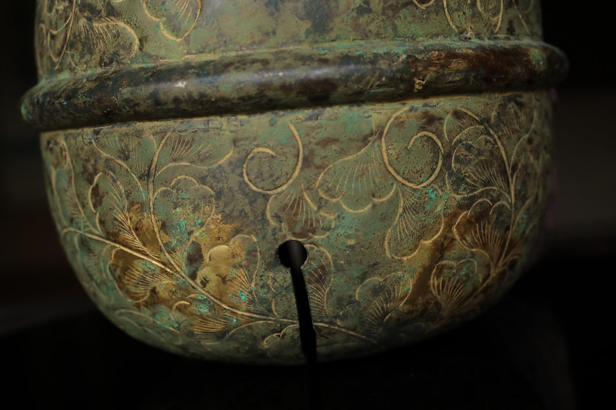 a close-up view of a decorative metal object, possibly a vase or a bowl, with intricate floral patterns and designs. The object appears to be made of a greenish-blue material and has a black hole in the center.
