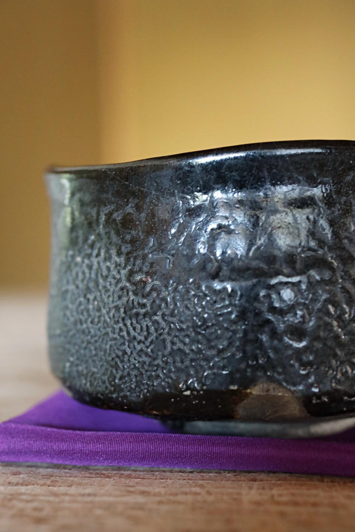 A close-up image of a black ceramic bowl with a textured surface, resting on a purple coaster on a wooden surface.