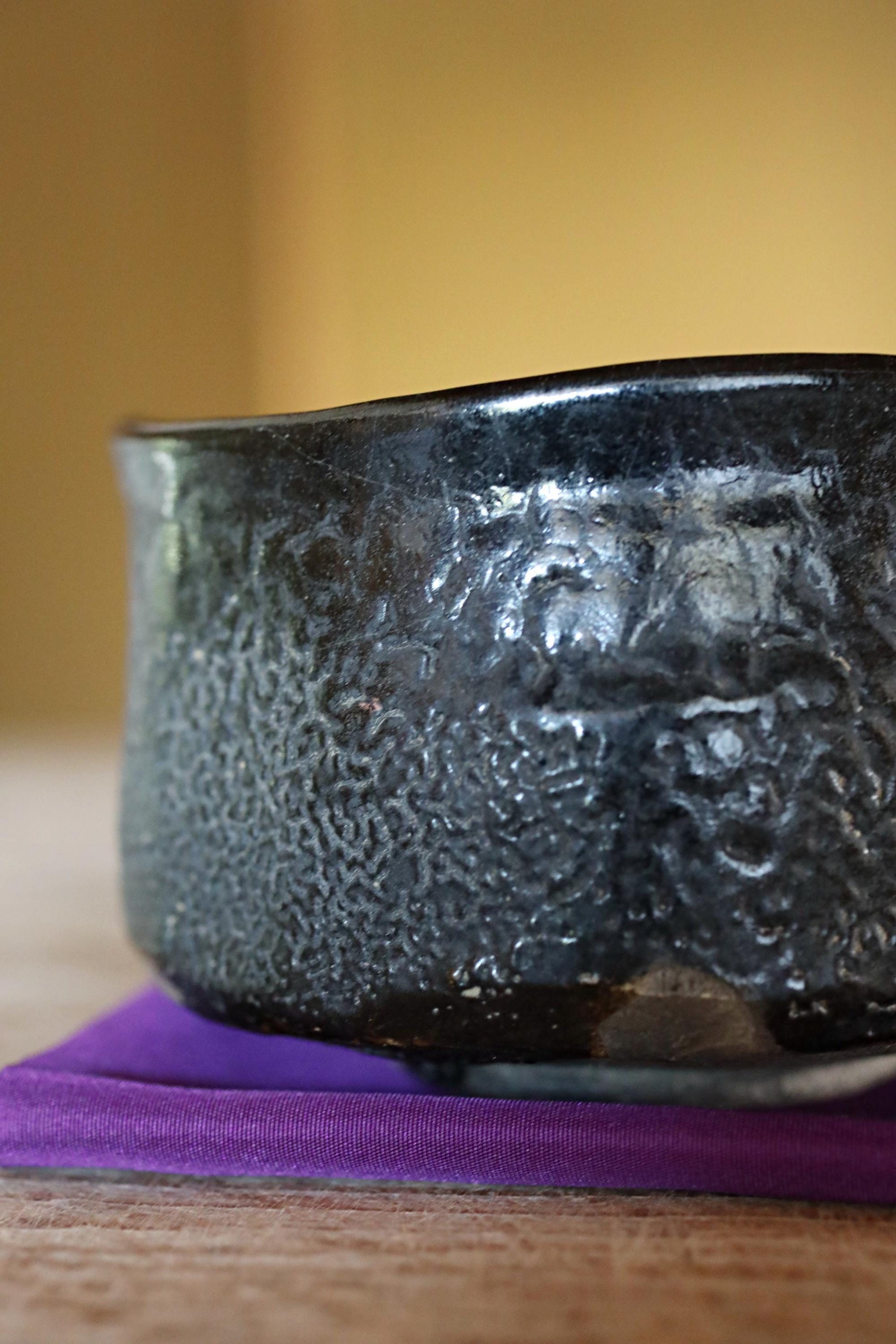 A close-up image of a black ceramic bowl with a textured surface, resting on a purple coaster on a wooden surface.