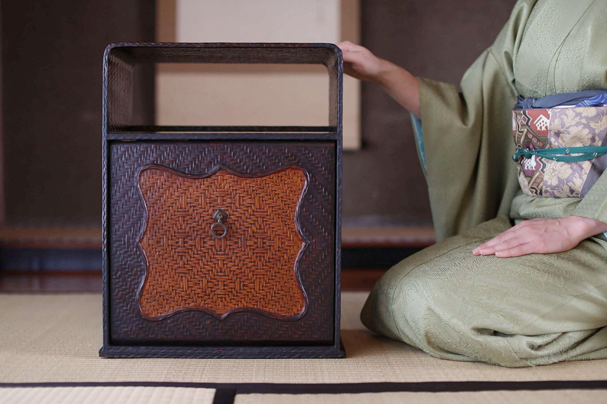 A person in traditional Japanese attire is kneeling next to a wooden cabinet with a woven design, possibly a storage unit or a decorative piece.