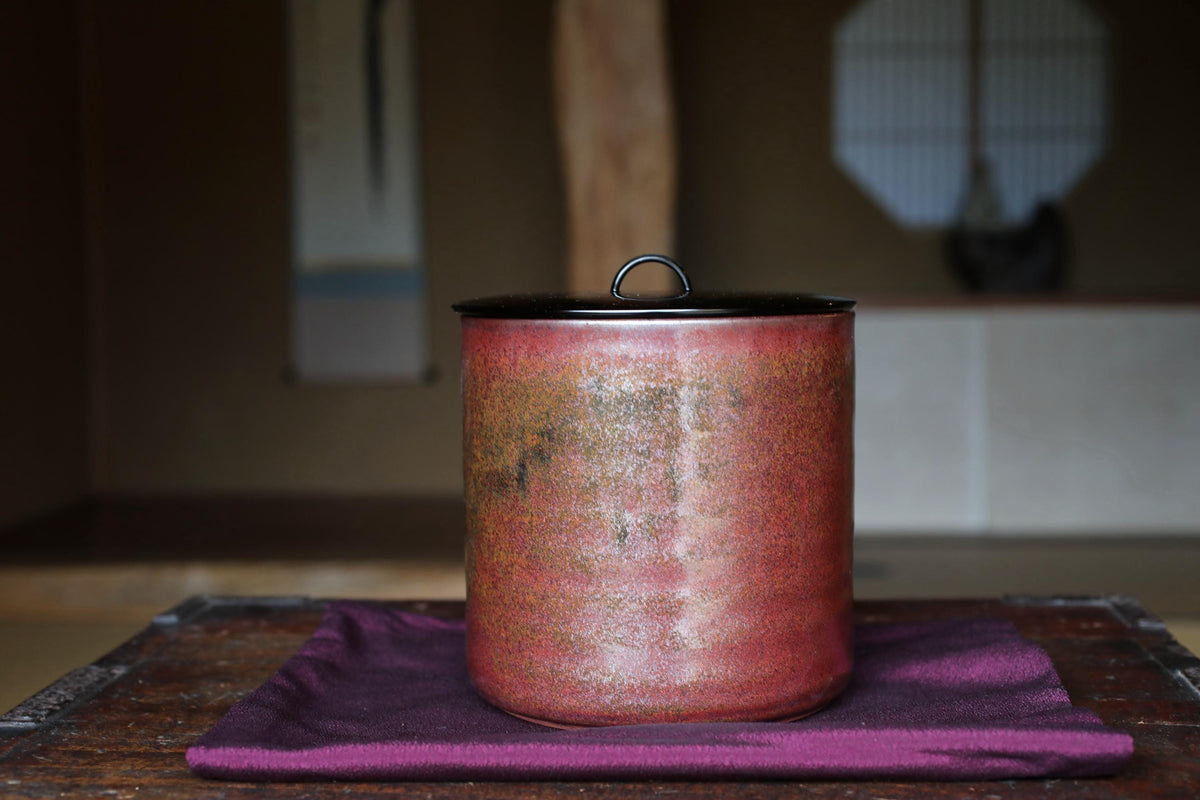 A red ceramic container with a black lid sits on a purple cloth on a wooden surface.