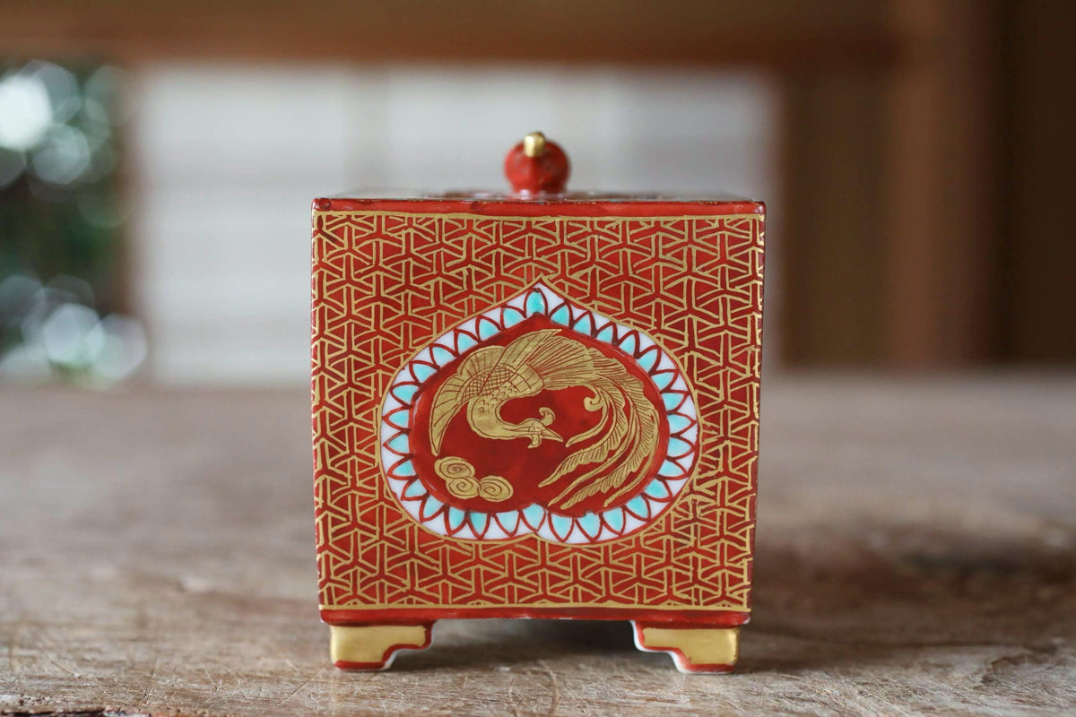 A red and gold decorative box with a gold finial and a gold bird design on the front, placed on a wooden surface.