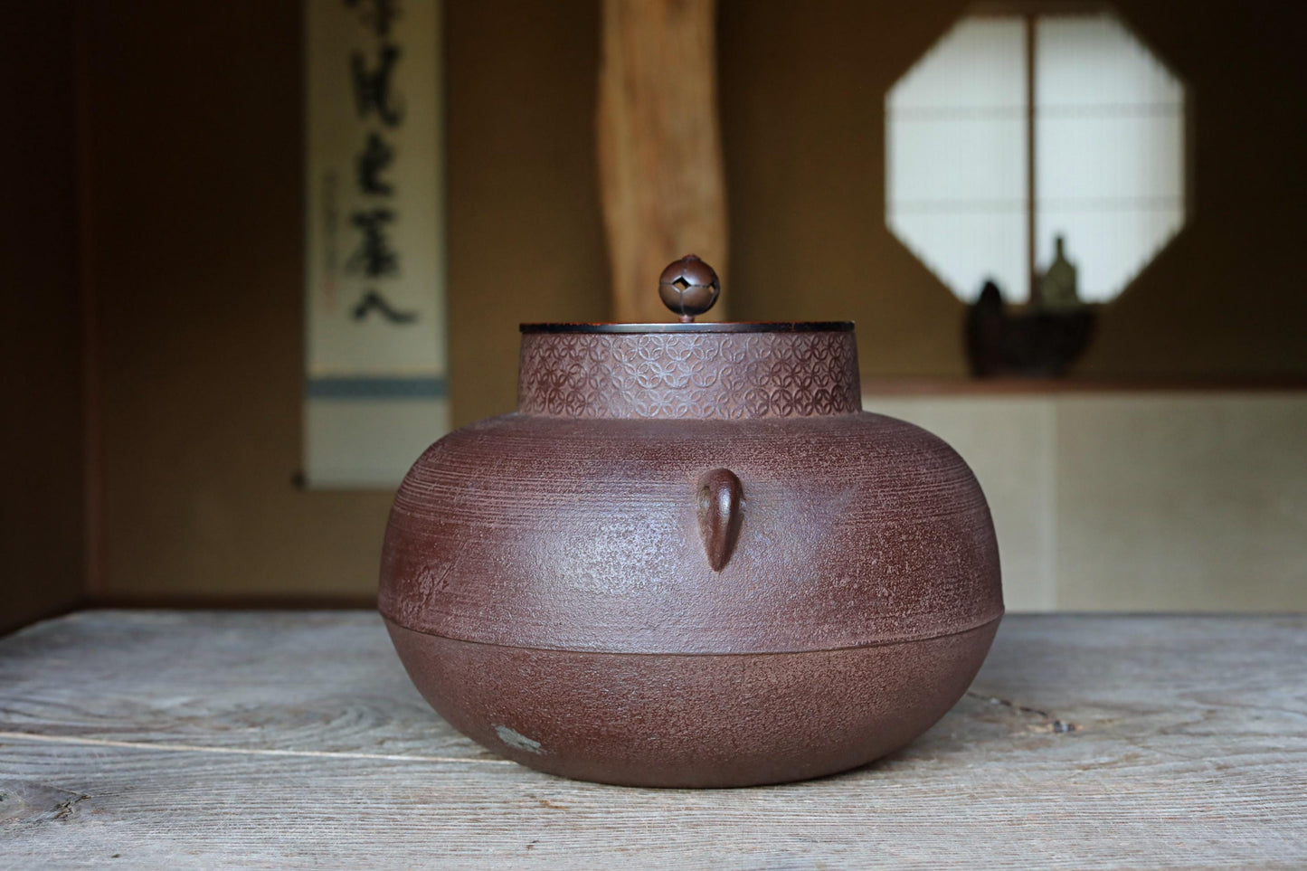 A brown ceramic teapot with a lid and spout sits on a wooden surface, with a window and a wooden beam in the background.