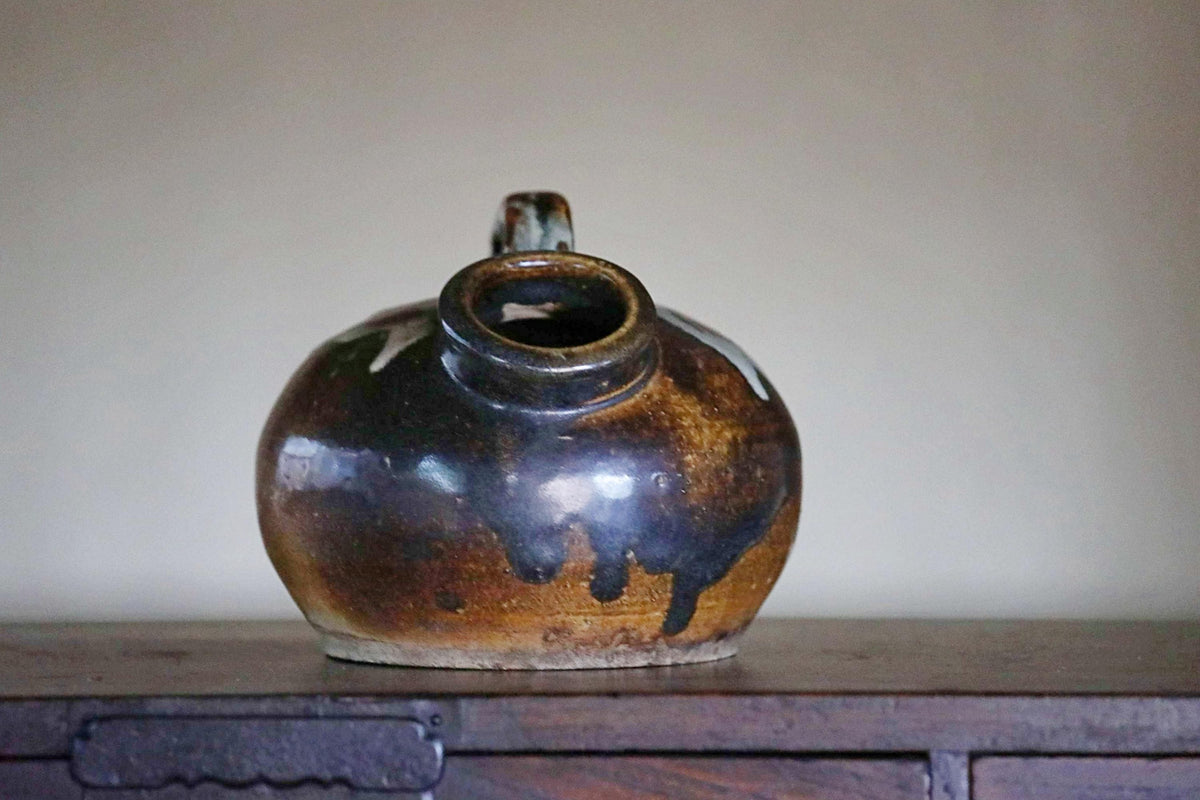 A ceramic pitcher with a dark brown glaze sits on a wooden surface against a plain background.