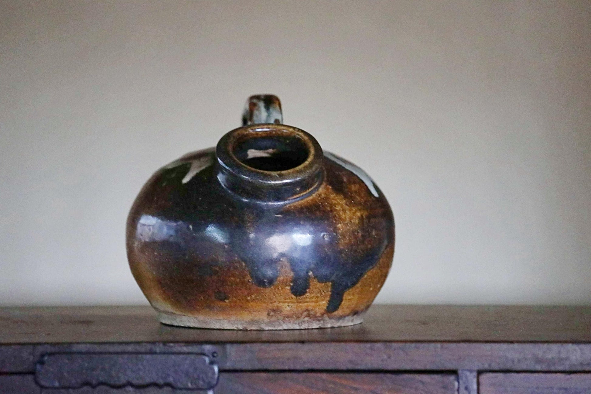 A ceramic pitcher with a dark brown glaze sits on a wooden surface against a plain background.