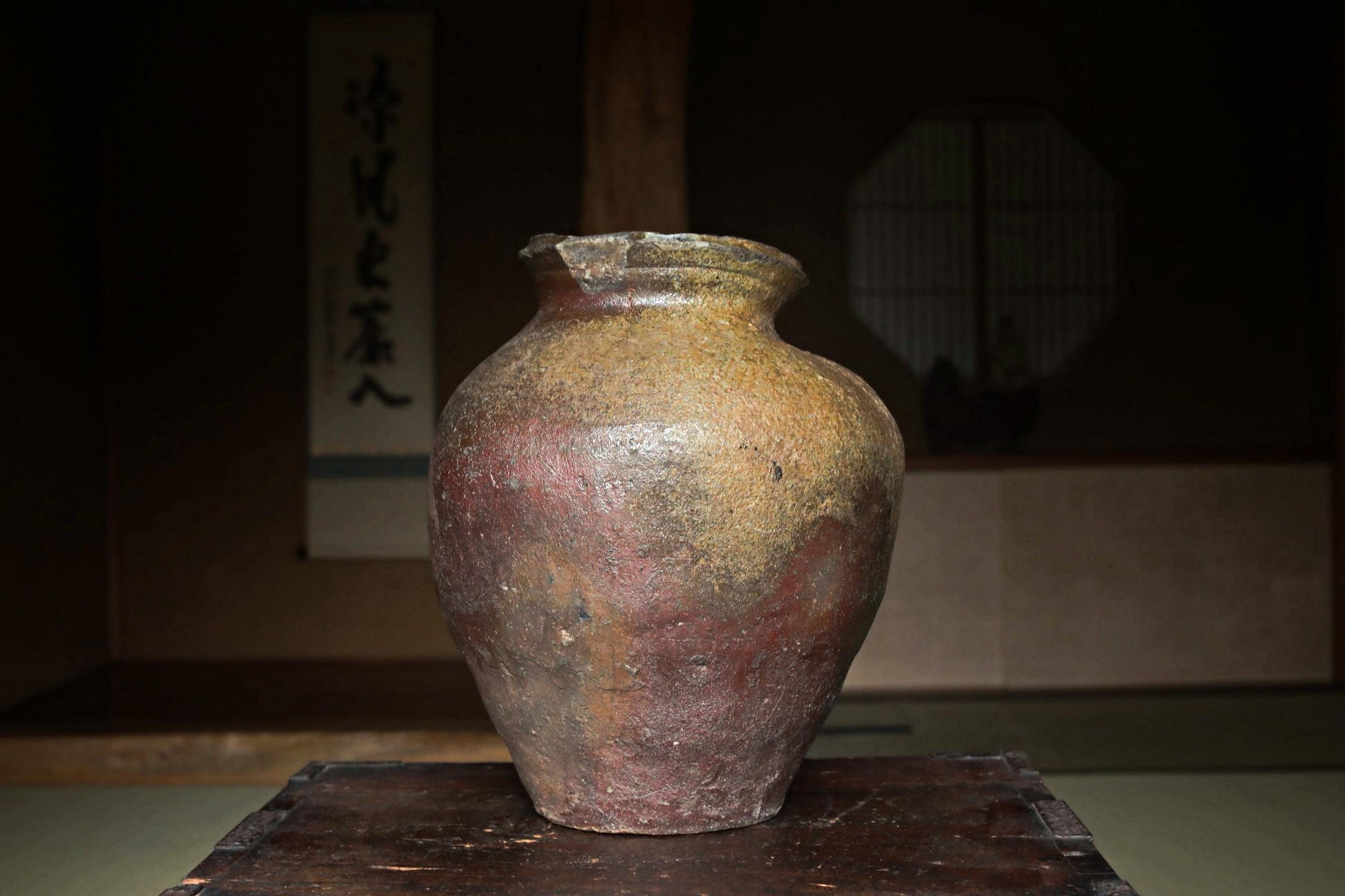 An old, weathered ceramic vase with a reddish-brown hue sits on a wooden surface, with a traditional Japanese-style room in the background.