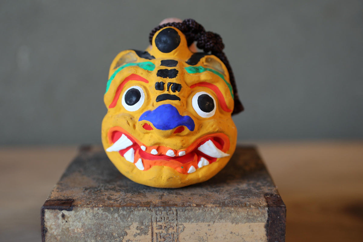 A colorful, painted mask with exaggerated features, such as large eyes, a prominent nose, and sharp teeth, is displayed on a wooden surface.