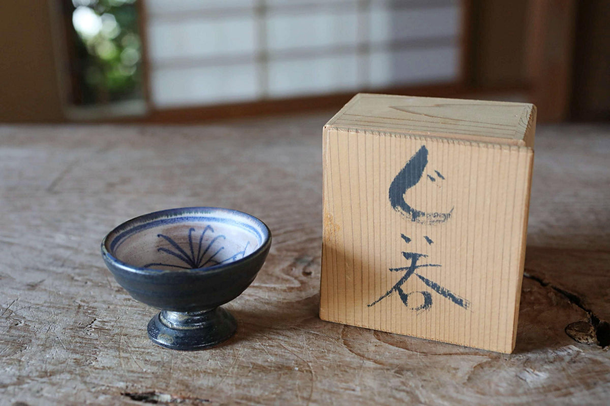 a small blue bowl with a decorative design on it, placed next to a wooden box with Japanese characters on it, both sitting on a wooden surface.