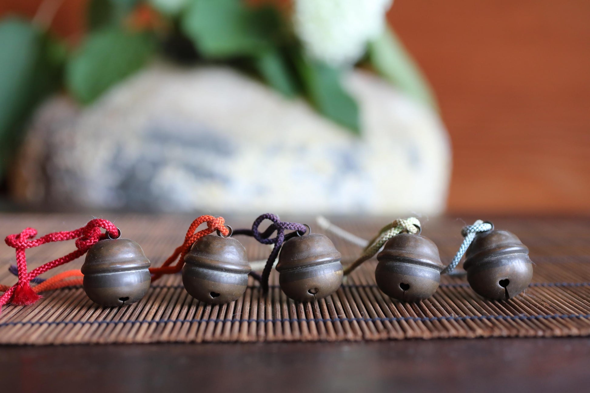 A row of five small, round metal bells with colorful strings hanging from them, arranged on a bamboo mat.