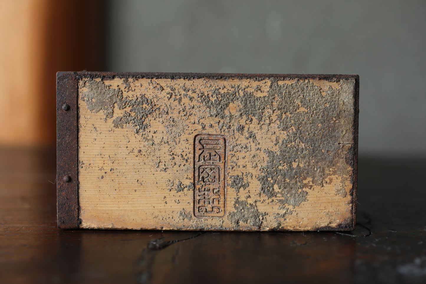 An old, weathered wooden object with a faded, distressed appearance, featuring a central, embossed symbol or text.