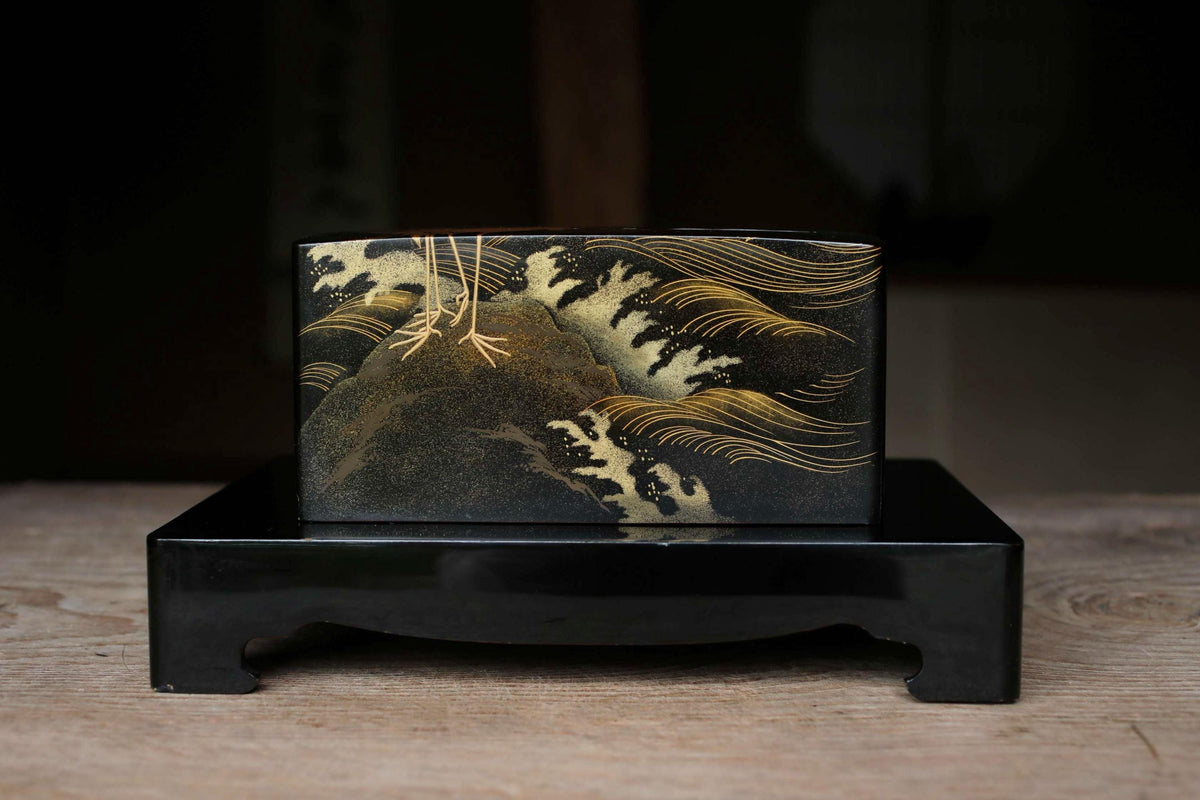 A black rectangular box with a gold and black Japanese-style design depicting waves and mountains on its surface, placed on a wooden surface.