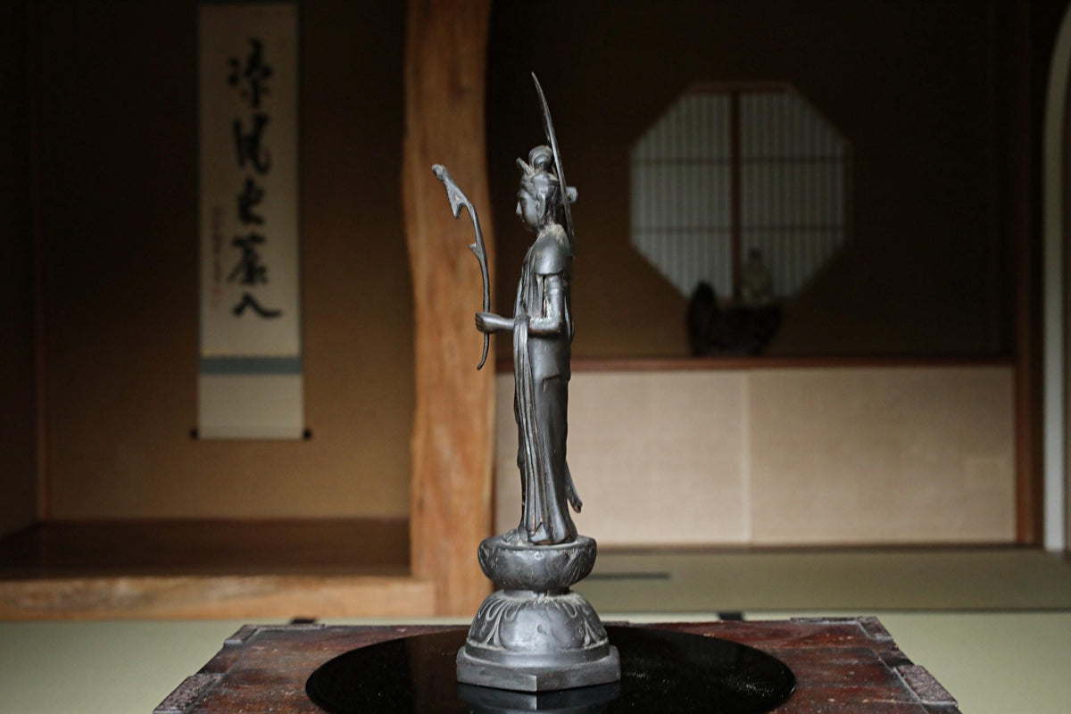 A silver statue of a deity stands on a wooden base in a dimly lit room with a traditional Japanese-style window and wall.
