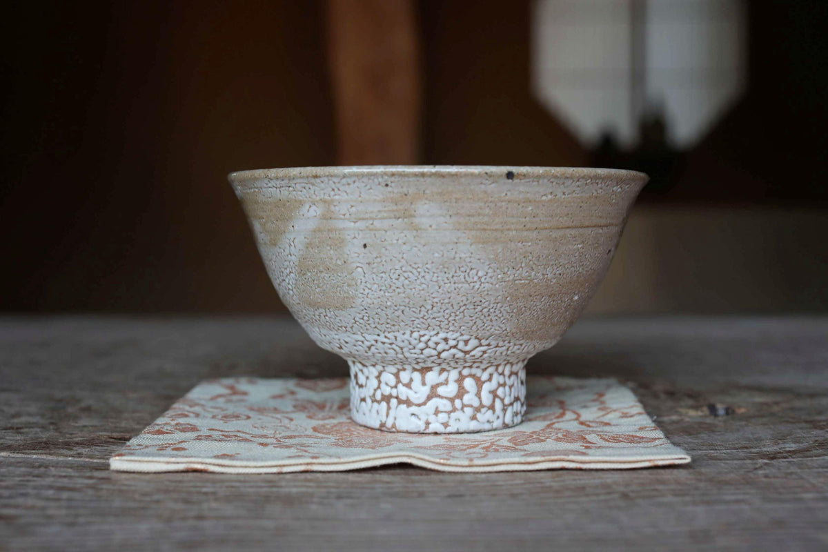 A white ceramic bowl with a speckled texture sits on a white and orange floral napkin on a wooden table.