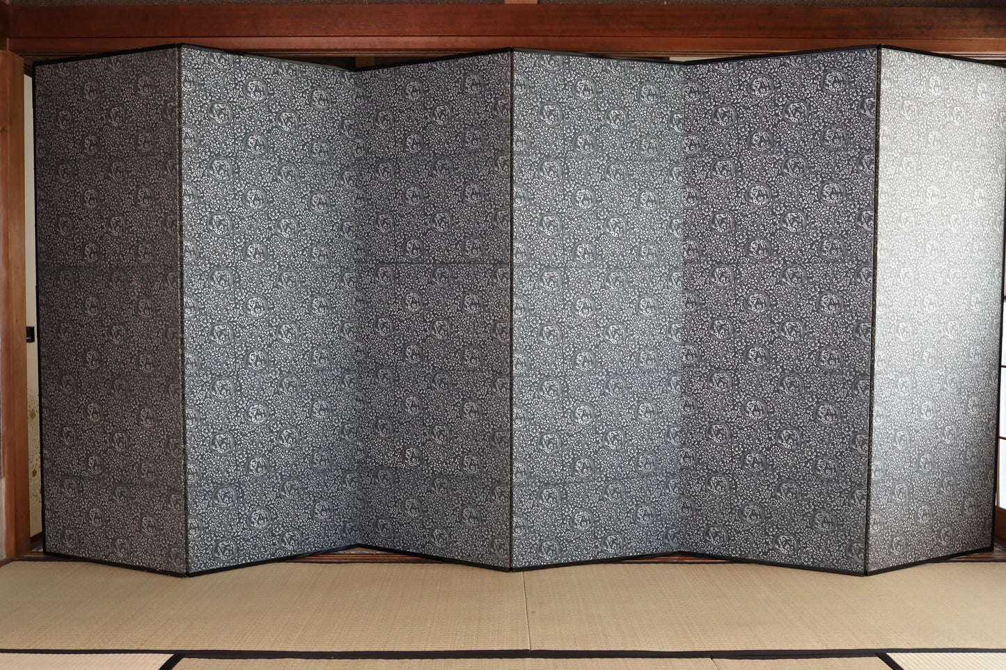a large, multi-paneled folding screen with a black and white patterned design. The screen is folded in half, creating a vertical structure that occupies a significant portion of the frame. The floor beneath the screen is covered with a beige mat, which contrasts with the dark color of the screen. The overall composition of the image is minimalistic and focused on the folding screen and its surroundings.