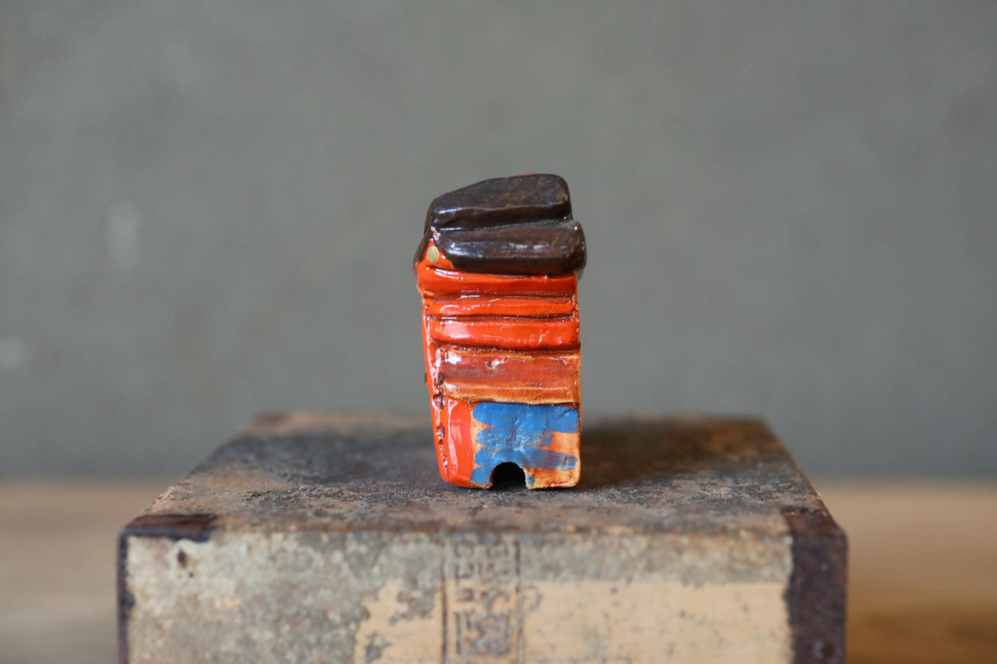A small, colorful ceramic sculpture with a dark, textured surface sits on a wooden surface against a gray background.