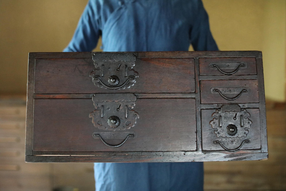 A person is holding a large wooden chest with multiple drawers and intricate metal handles.