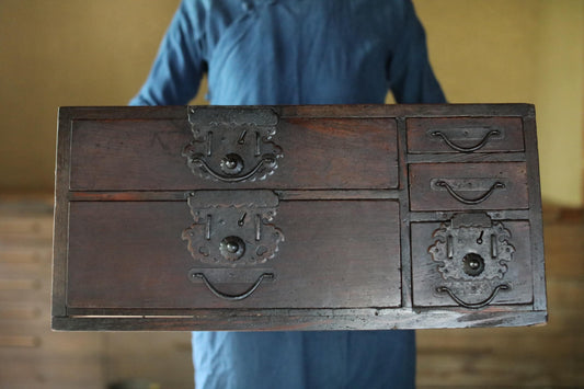 A person is holding a large wooden chest with multiple drawers and intricate metal handles.