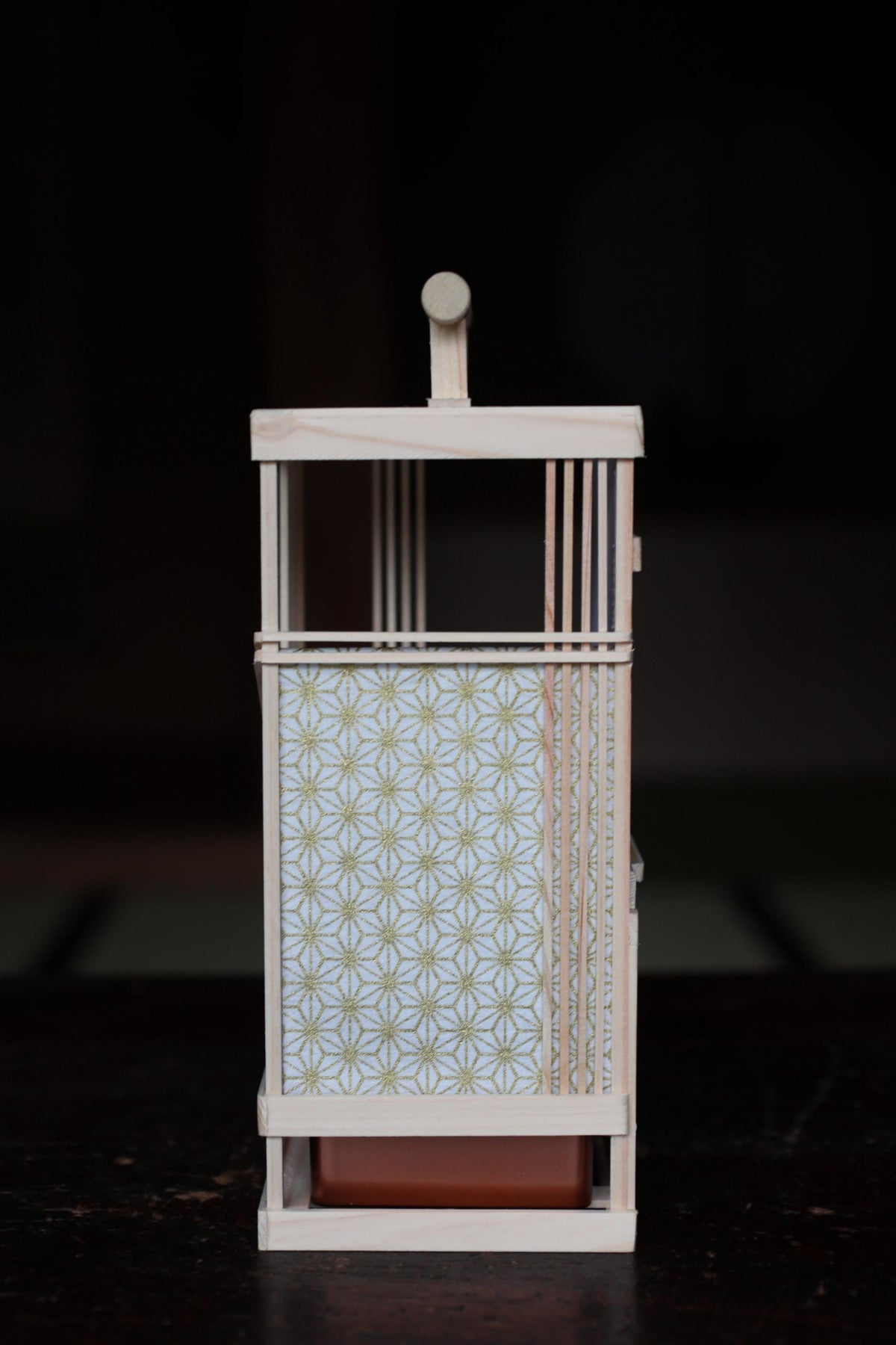 A wooden lantern with a white and gold patterned design on its front, featuring a wooden handle and a small red base.