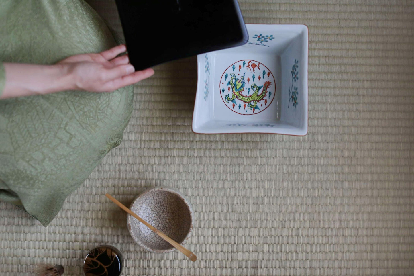 a person's hand holding a black object, which appears to be a tablet or electronic device, next to a white ceramic bowl with a dragon design. The bowl is placed on a woven mat or rug, and there are other small items, such as a wooden spoon, a small bowl, and a cup, scattered around the scene.