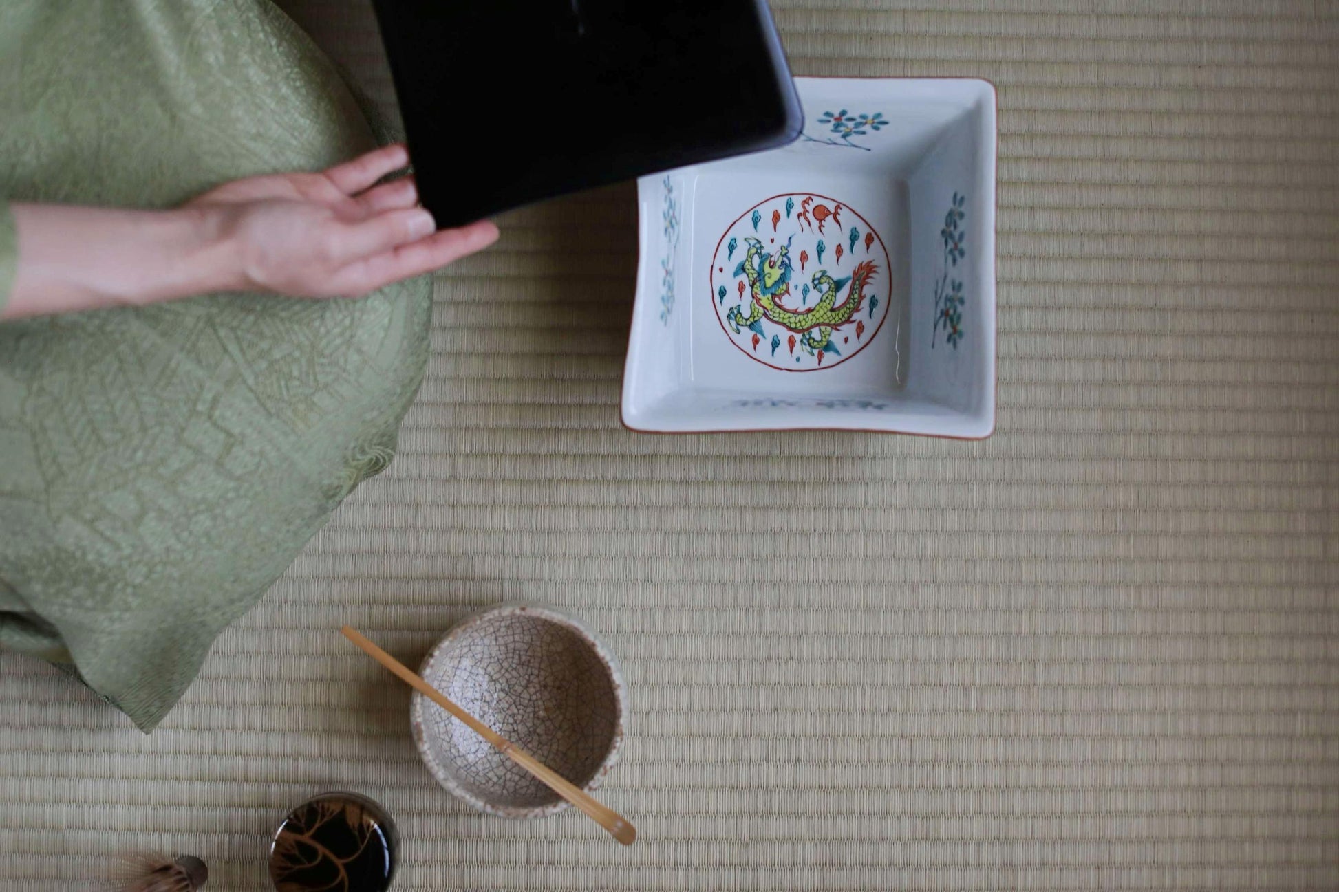 a person's hand holding a black object, which appears to be a tablet or electronic device, next to a white ceramic bowl with a dragon design. The bowl is placed on a woven mat or rug, and there are other small items, such as a wooden spoon, a small bowl, and a cup, scattered around the scene.