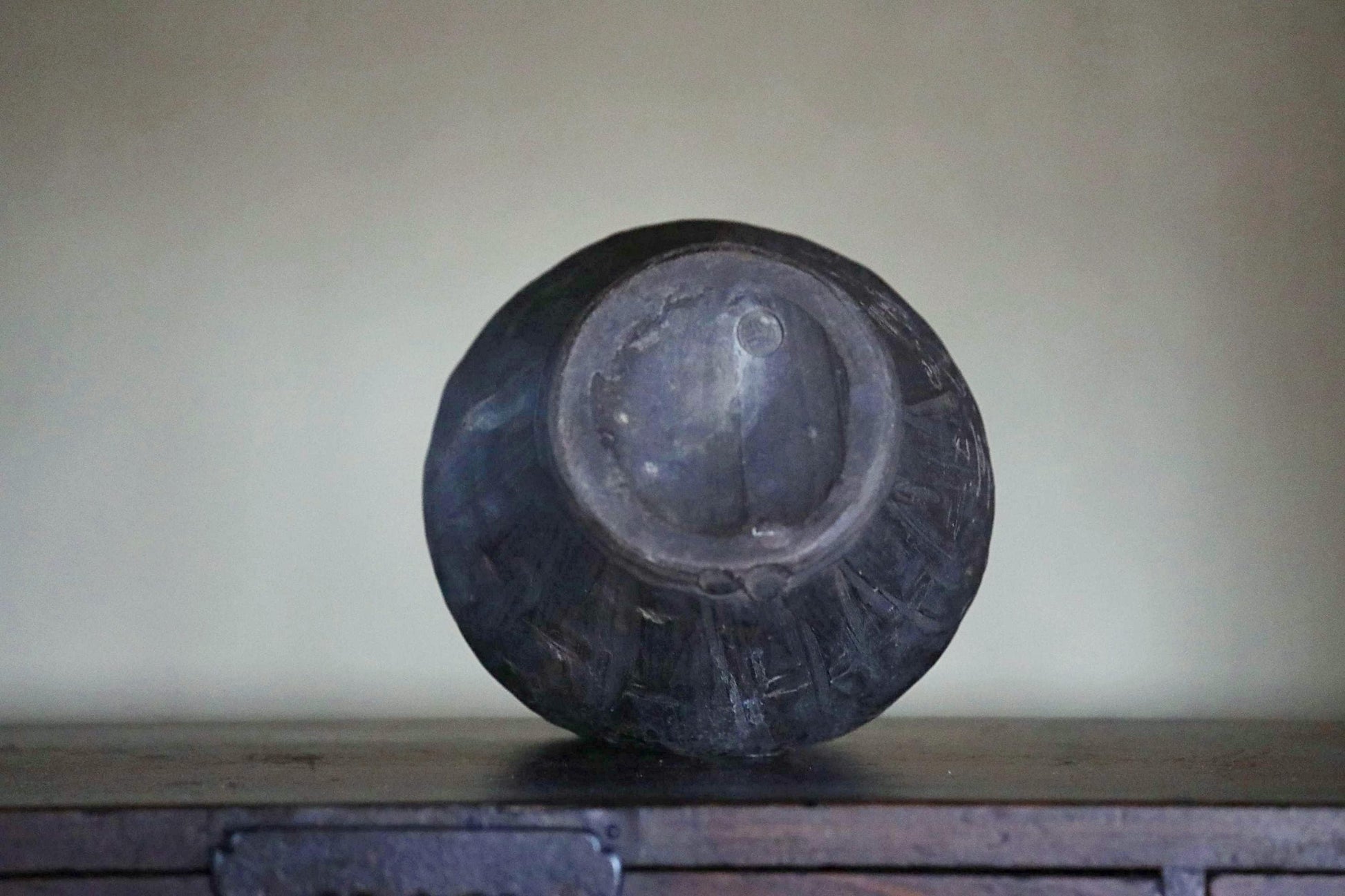 A dark, round vase sits on a wooden surface against a plain background.