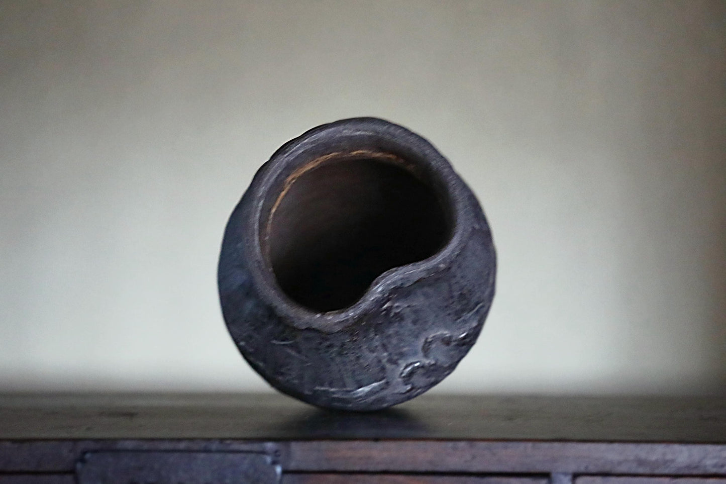 A dark, textured ceramic bowl or vase with a large opening, sitting on a wooden surface against a plain background.