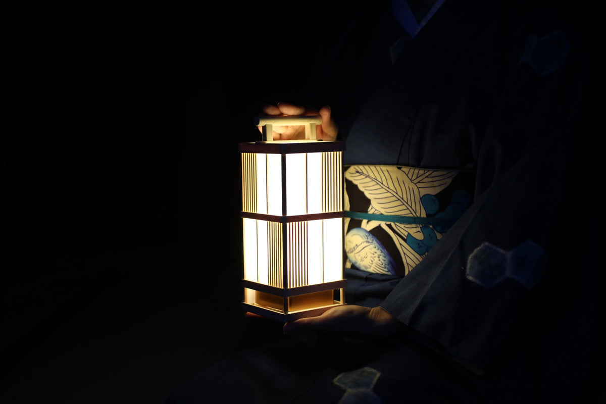 A person wearing a traditional Japanese kimono holds a lit lantern in a dark setting.