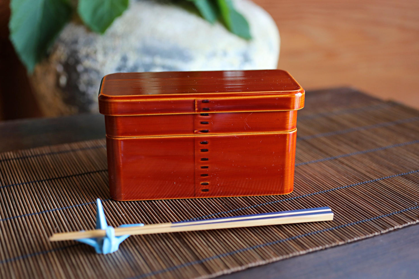 A red wooden box with a lid and a pair of chopsticks on a bamboo placemat.