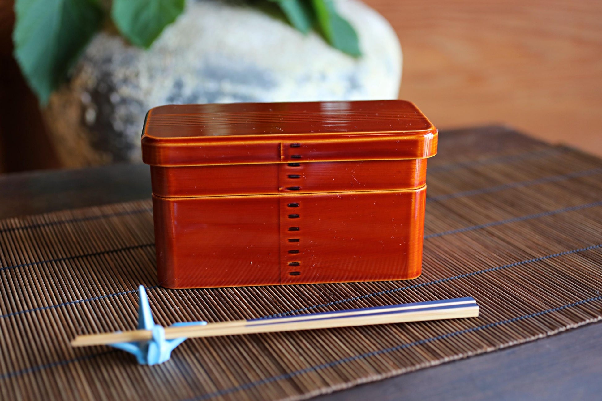 A red wooden box with a lid and a pair of chopsticks on a bamboo placemat.