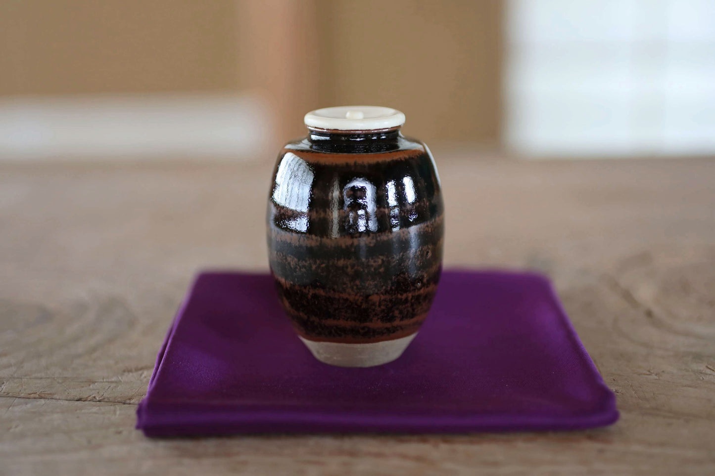 A small, round ceramic jar with a white lid sits on a purple cloth on a wooden surface.