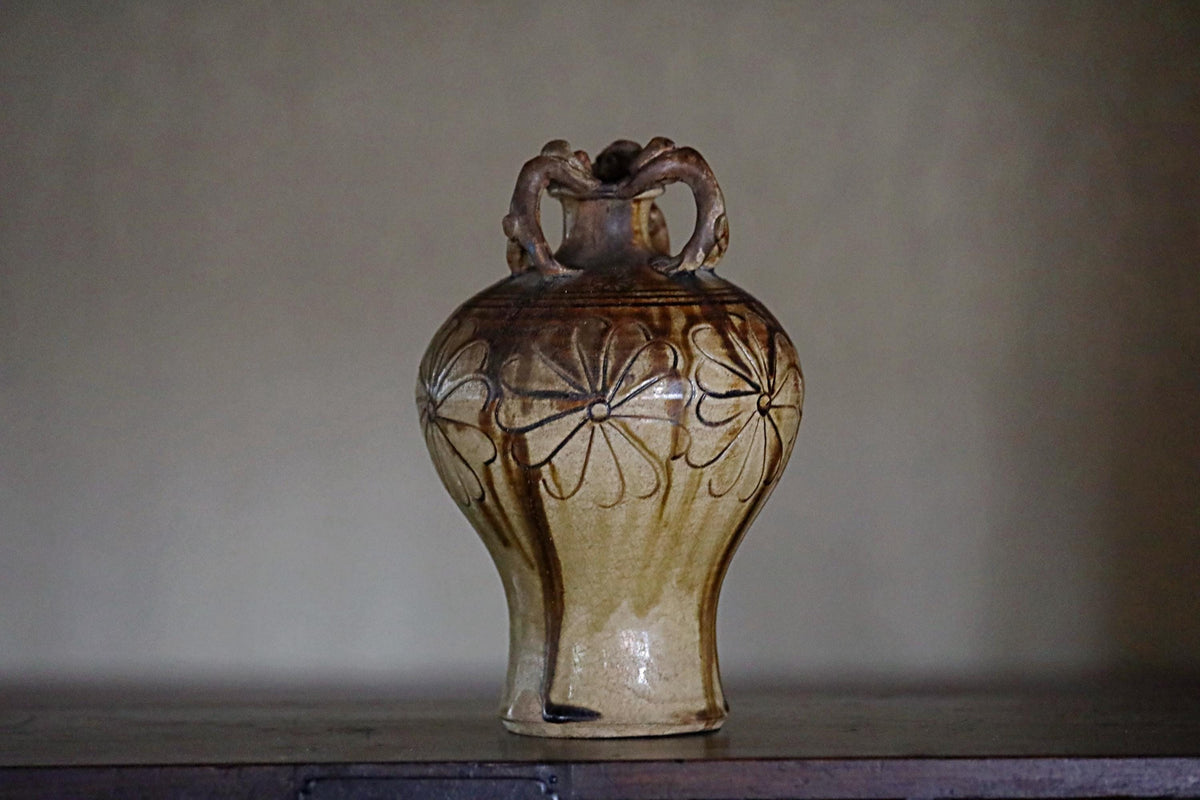 A ceramic vase with a handle and floral design on its body, sitting on a wooden surface against a plain background.