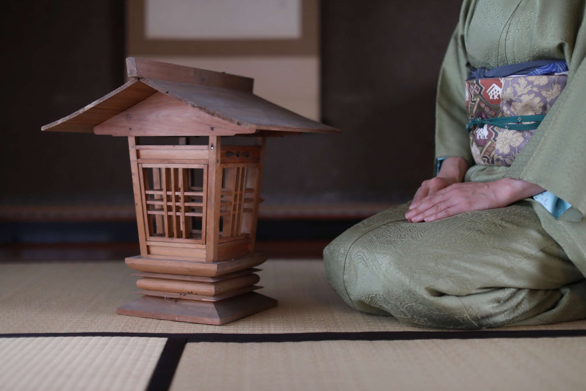 A person in a green kimono is kneeling on a tatami mat, with a small wooden lantern or lantern-like structure next to them.